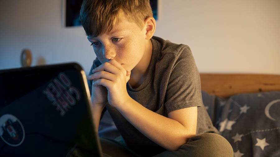 A young boy in a bedroom, at a laptop, with their head in their hands