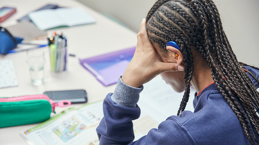 A mixed-race boy with braided hair and a hearing aid, is looking at school work, he seems stressed