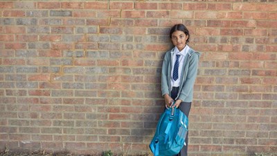 A young girl in school uniform standing in front of a brick wall while smiling.