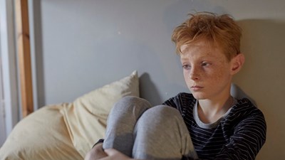 A young boy sitting on his bed looking sad.