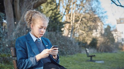 A teenage girl in school uniform on her mobile while sitting on a park bench.