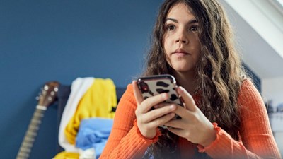 A teenage girl on her mobile in her bedroom while staring into the distance.