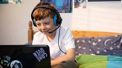 A young boy wearing headphones while using a laptop on a bed.