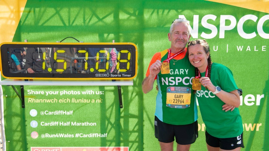 Two NSPCC Cardiff Half runners, holding up their finisher medals, pose next to the finish time clock