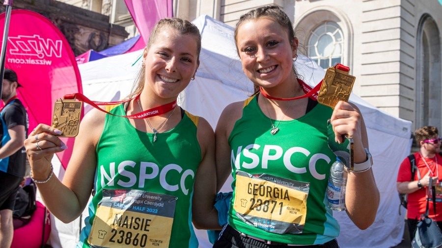 Two NSPCC Cardiff Half runners, standing outside, hold up their finisher medals