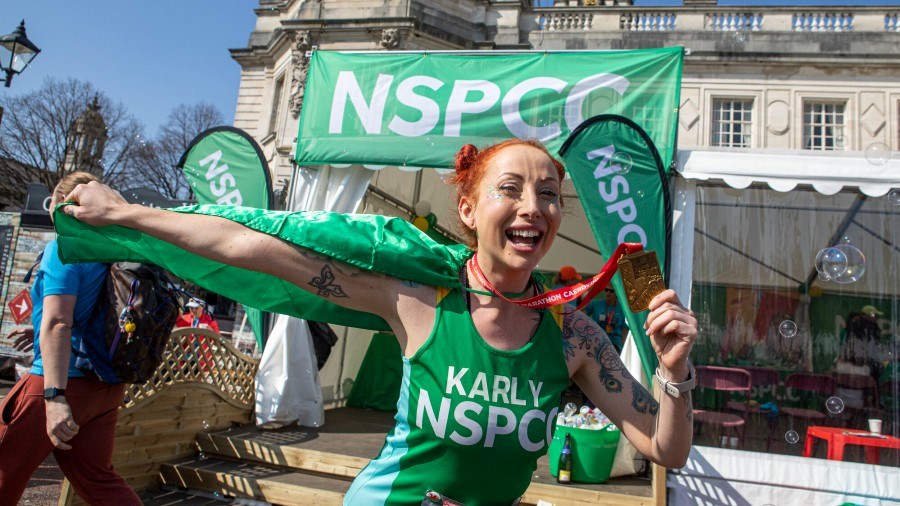 An NSPCC Cardiff Half runner, standing outside, holds up both their green cape and finisher medal