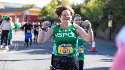 A woman in a green NSPCC running vest running with her arms in the air.