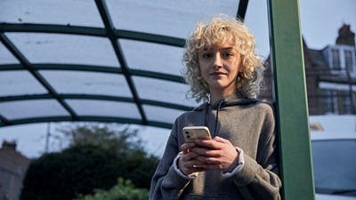 A teenage girl using her phone outside a bus stop while smiling into the camera.