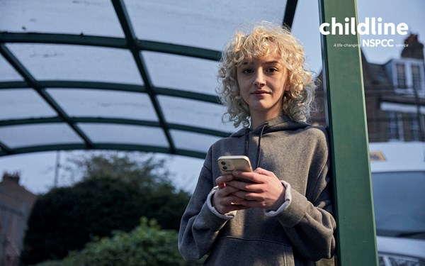 A teenage girl using her phone outside a bus stop while smiling into the camera. A white NSPCC and Childline logo lockup is layered into the top right corner of the image which says 'Childline: a life-saving NSPCC service'.