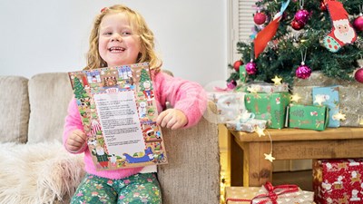 A young girl smiling while holding a Letter from Santa beside a Christmas tree.