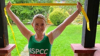 A woman in a green NSPCC running vest holding a yellow finish line ribbon in the air while smiling into the camera.