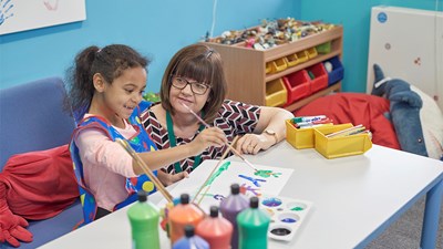 A young girl smiling while painting with a teacher at nursery.