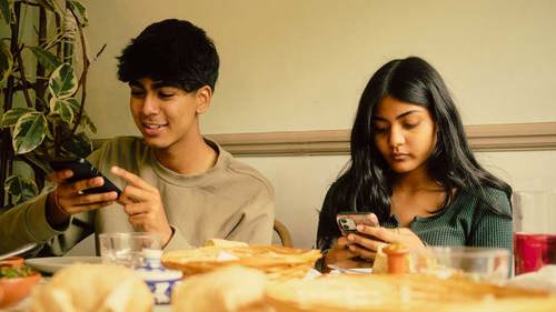 A teenage boy and teenage girl are sitting side by side at a table filled with food. They are both looking at their phones.