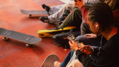Photograph of teenagers sat inside with skateboards, using their mobile phones. 