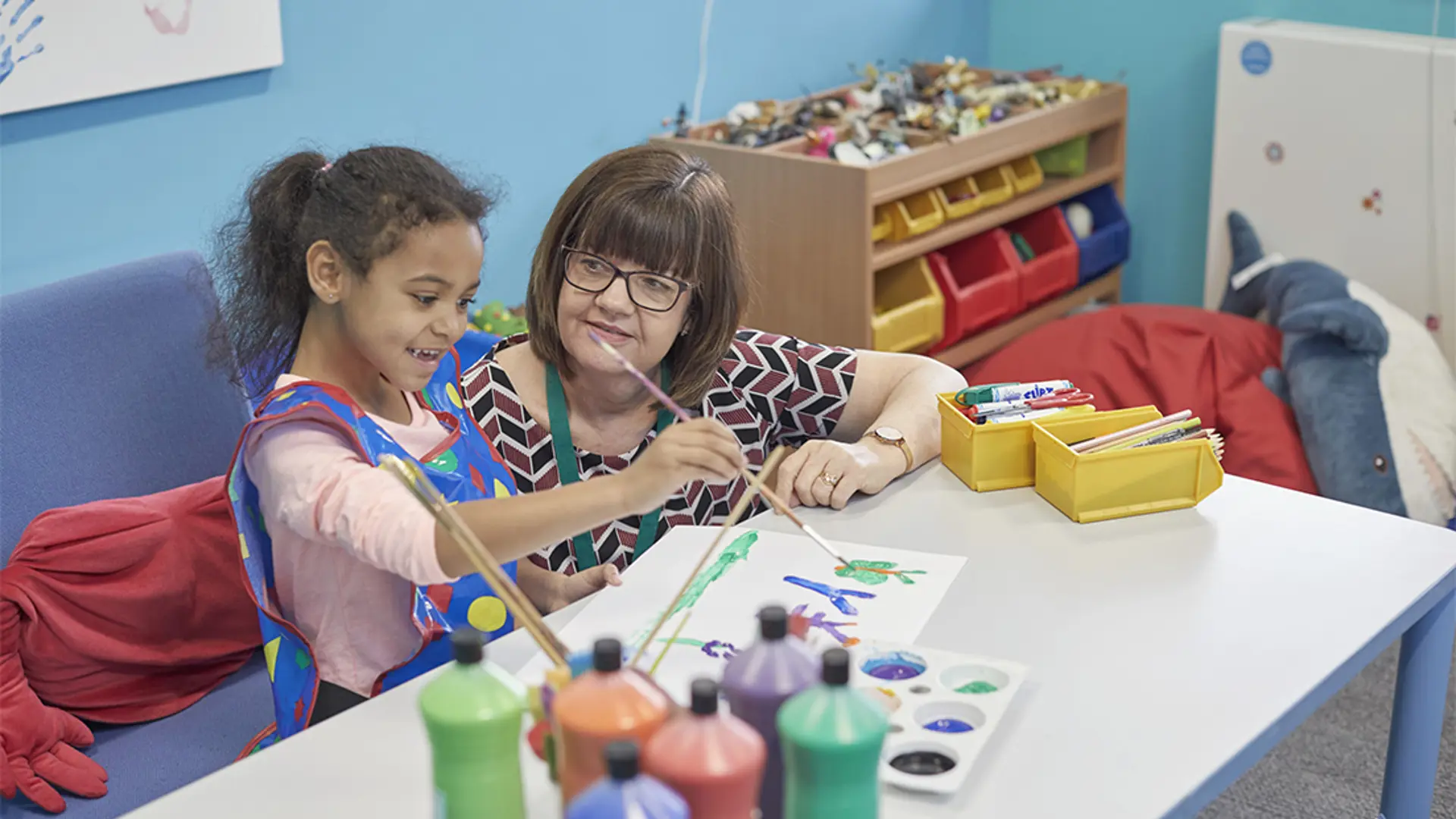 A young girl smiling while painting with a teacher at nursery.