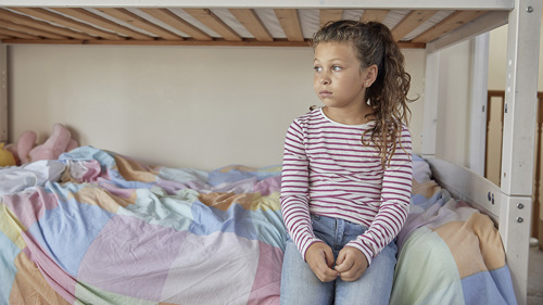 A young child sat on their bed while staring blankly into the distance.